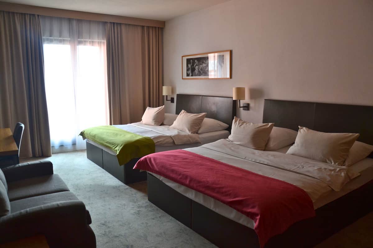 A hotel room with two beds, a sofa, a desk, and a large window with curtains.
