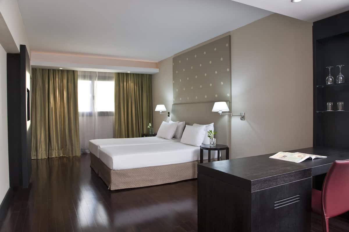 Une chambre d’hôtel avec un lit, un bureau et une chaise. Il y a une fenêtre avec des rideaux, une étagère fixée au mur avec des verres, et une petite table avec une lampe.