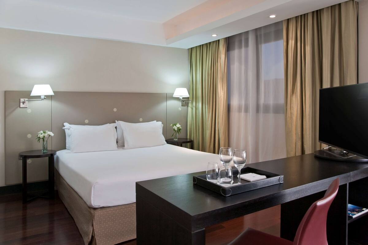 Une chambre d’hôtel avec un lit, des tables de chevet, un bureau avec un plateau de verres, une télévision et des rideaux.
