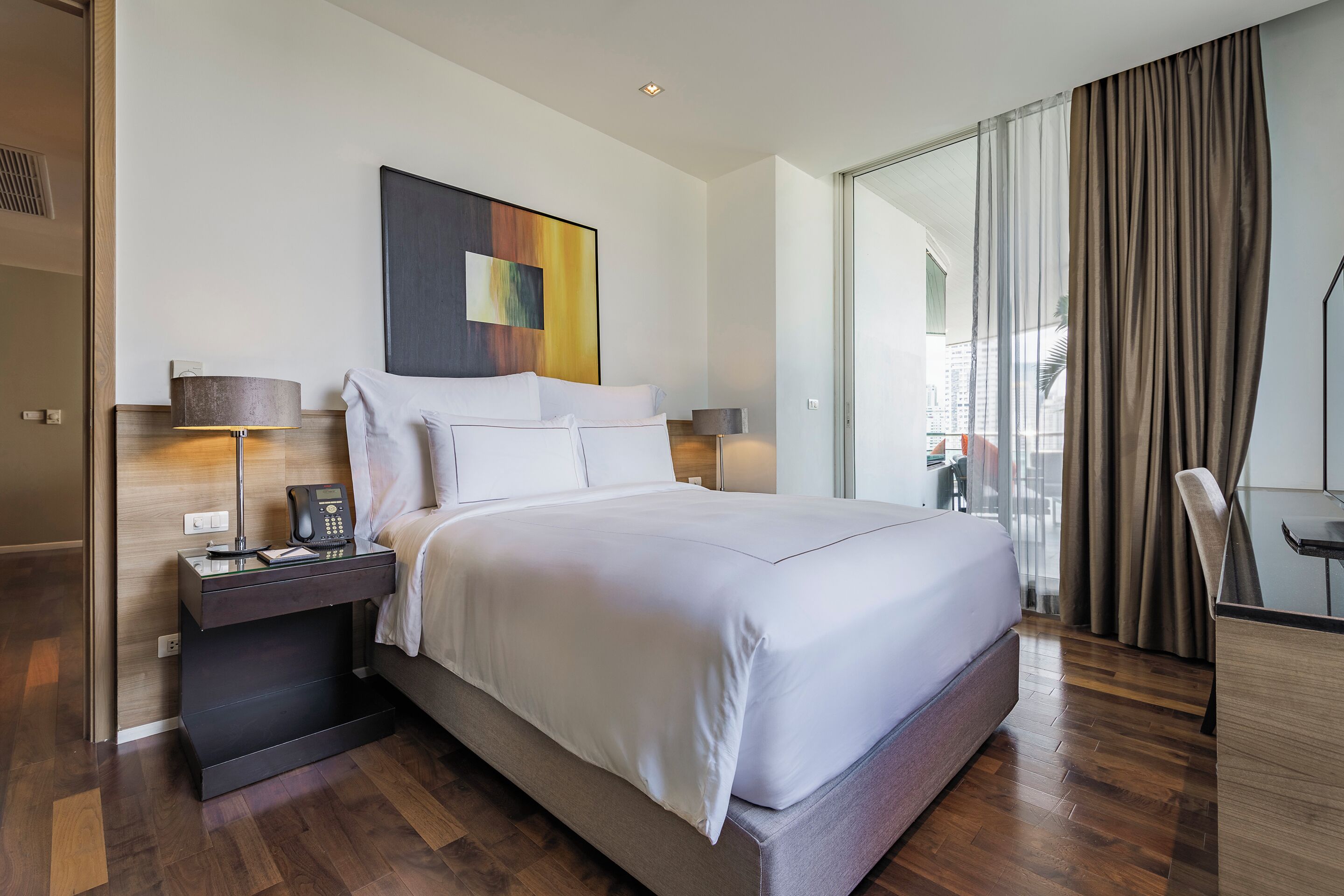 A modern hotel room with a large bed, bedside lamps, a telephone, and a view of the city through a large window.