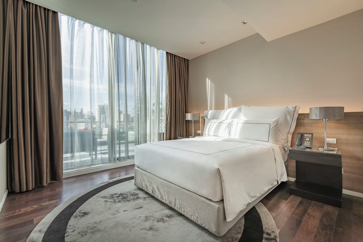 Moderni hotellihuone, jossa on suuri sänky, yöpöytä lampun kanssa ja näkymä kaupunkiin lattiasta kattoon ulottuvien ikkunoiden kautta.