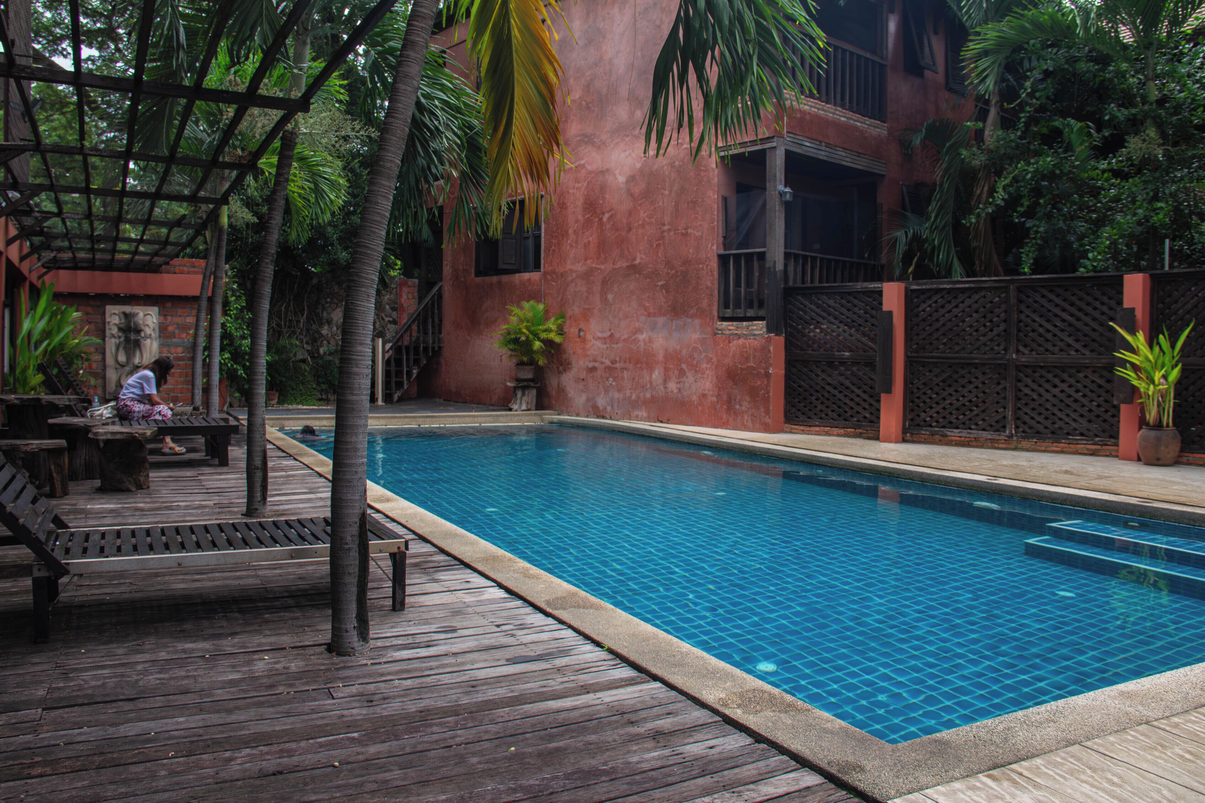 A swimming pool with a wooden deck, surrounded by palm trees and a brick building.