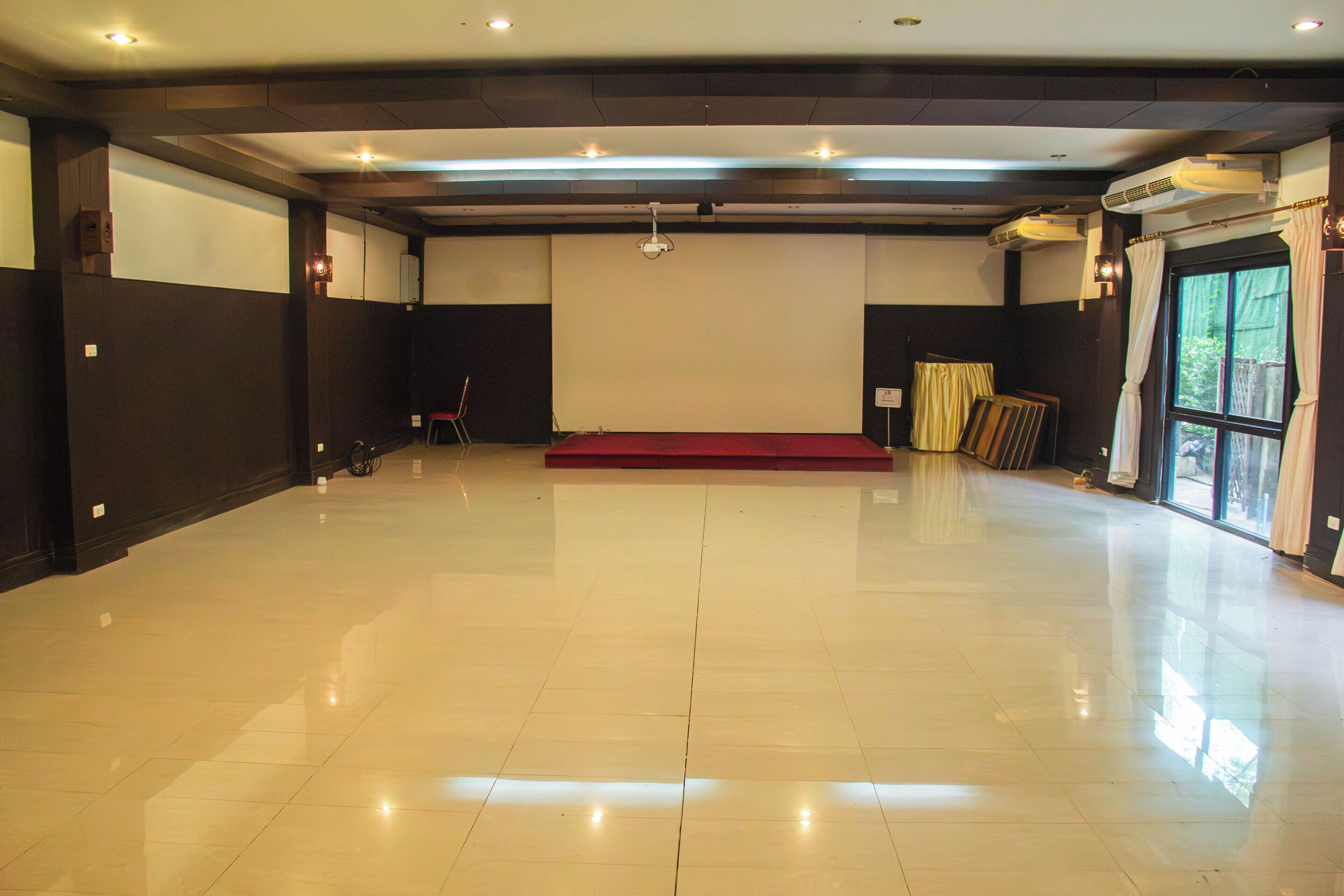 A spacious room with a polished floor, a stage with a red carpet, and a large window with curtains.