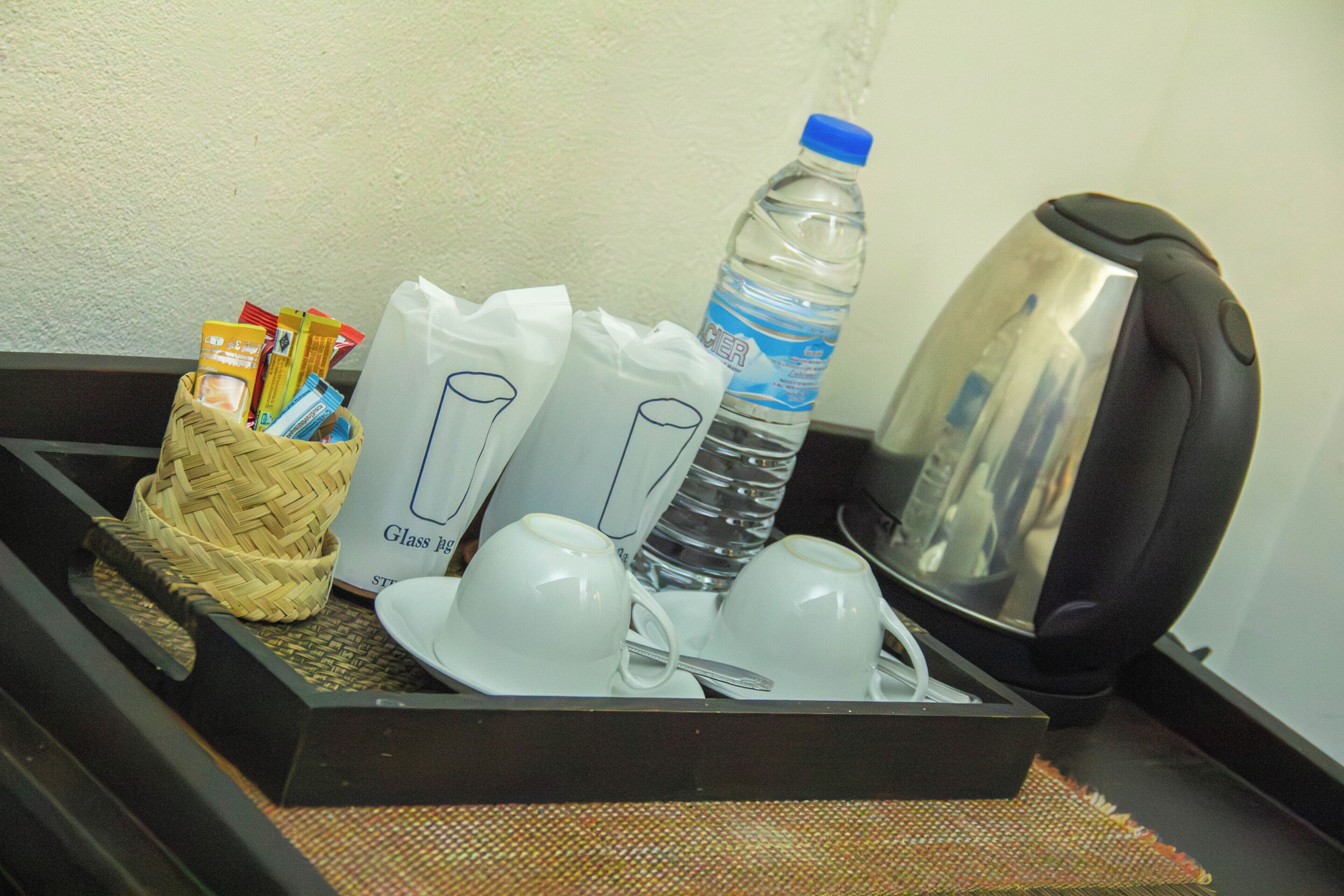A tray with a water bottle, a kettle, a woven basket with toiletries, and a glass pitcher.