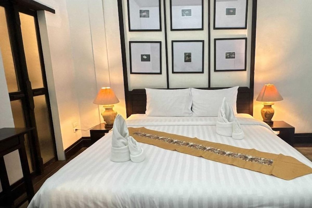 A hotel room with a large bed, two bedside lamps, a headboard with framed pictures, and a decorative towel on the bed.