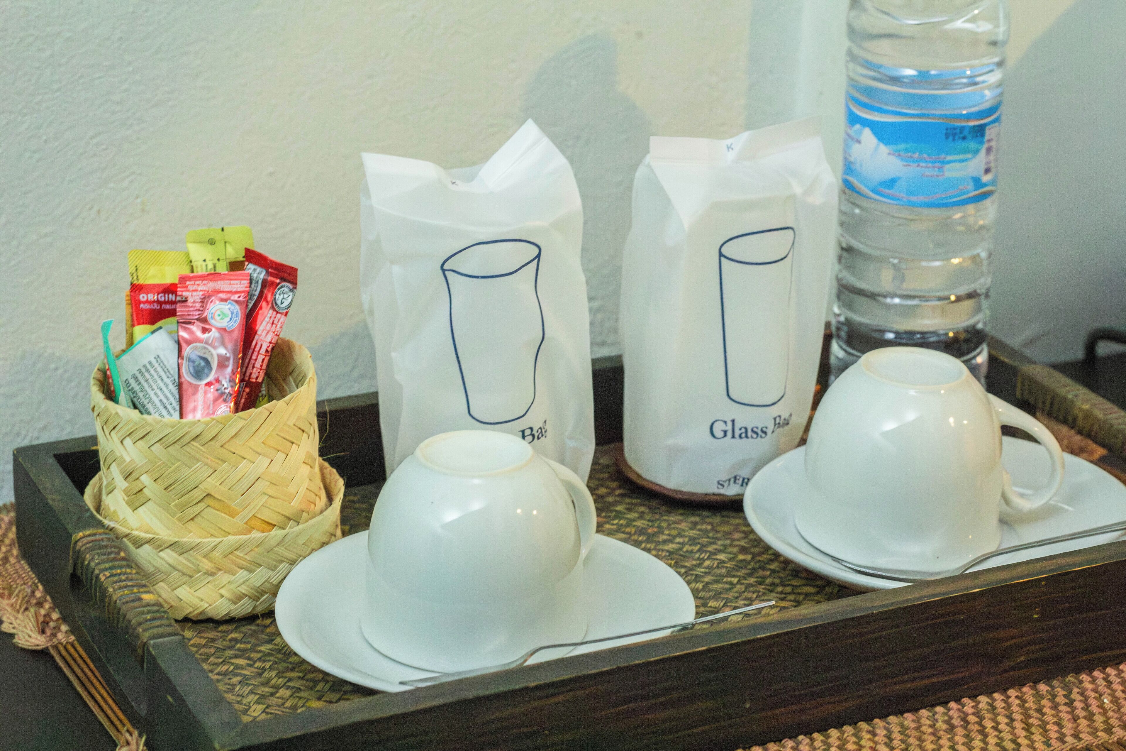 A tray with two white cups and saucers, a woven basket with packets, and a water bottle.