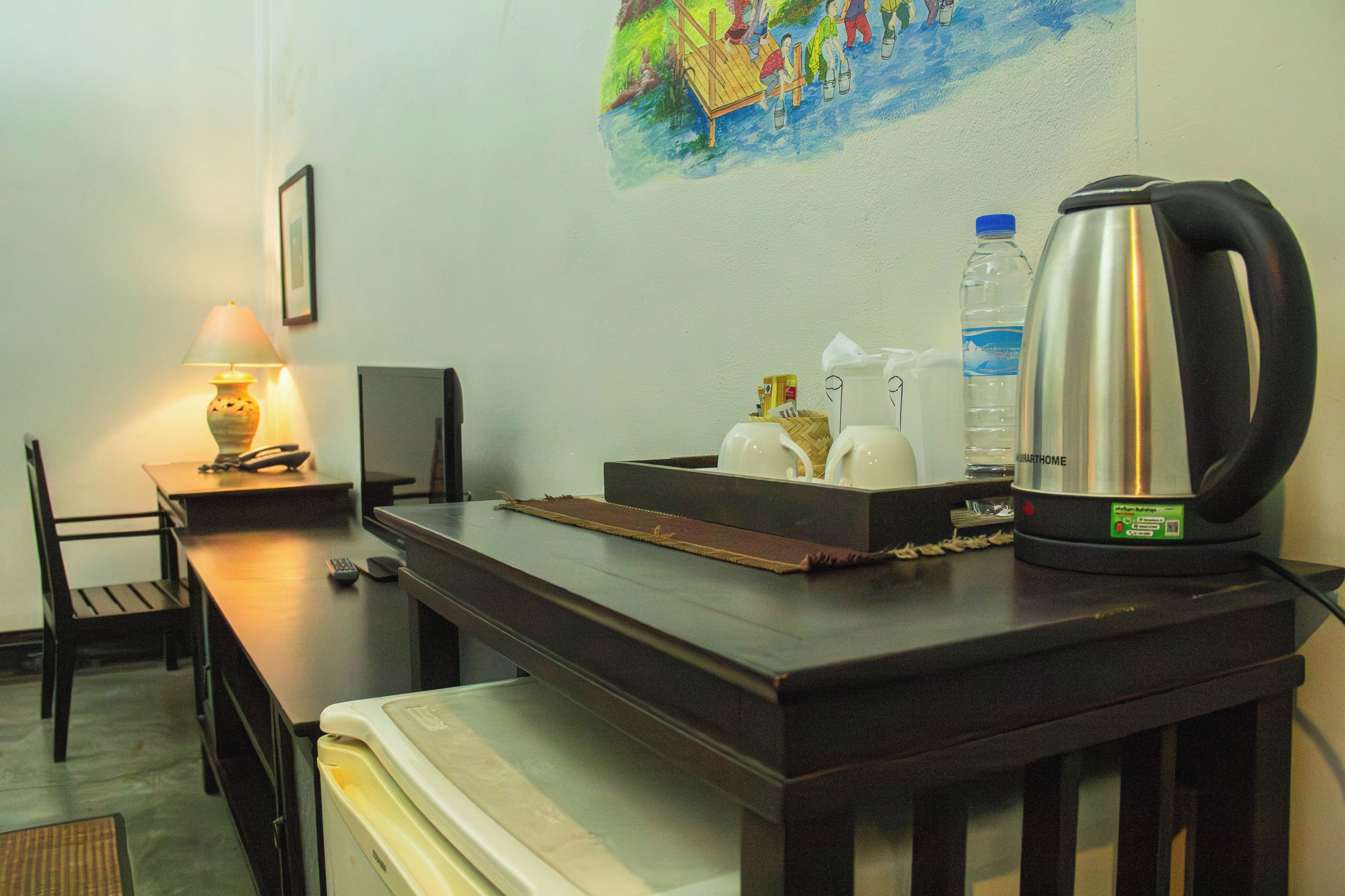 A hotel room with a desk, a lamp, a painting on the wall, a water bottle, a kettle, and a remote control.