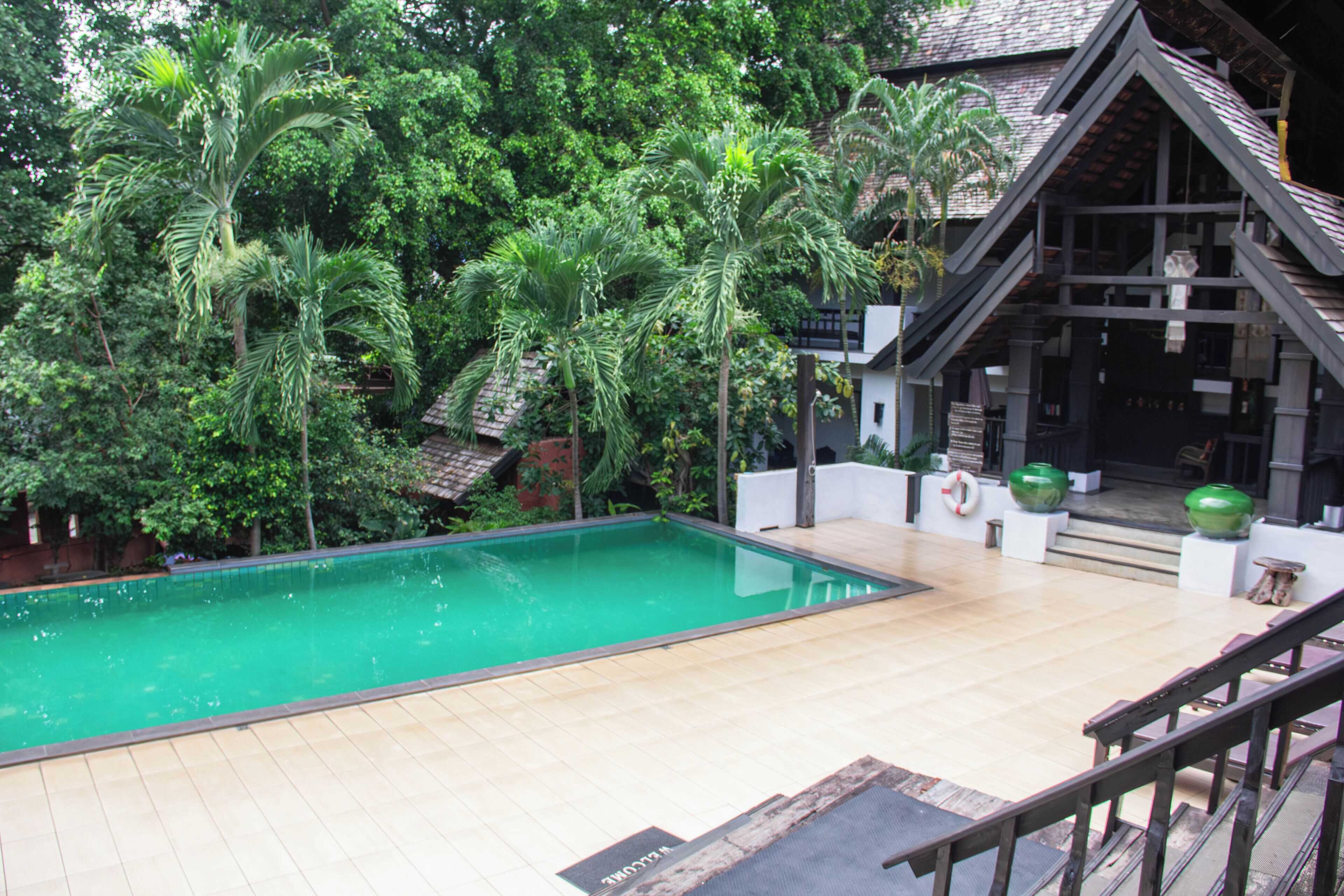 A swimming pool with a tiled deck, surrounded by lush greenery and a building with a sloped roof.