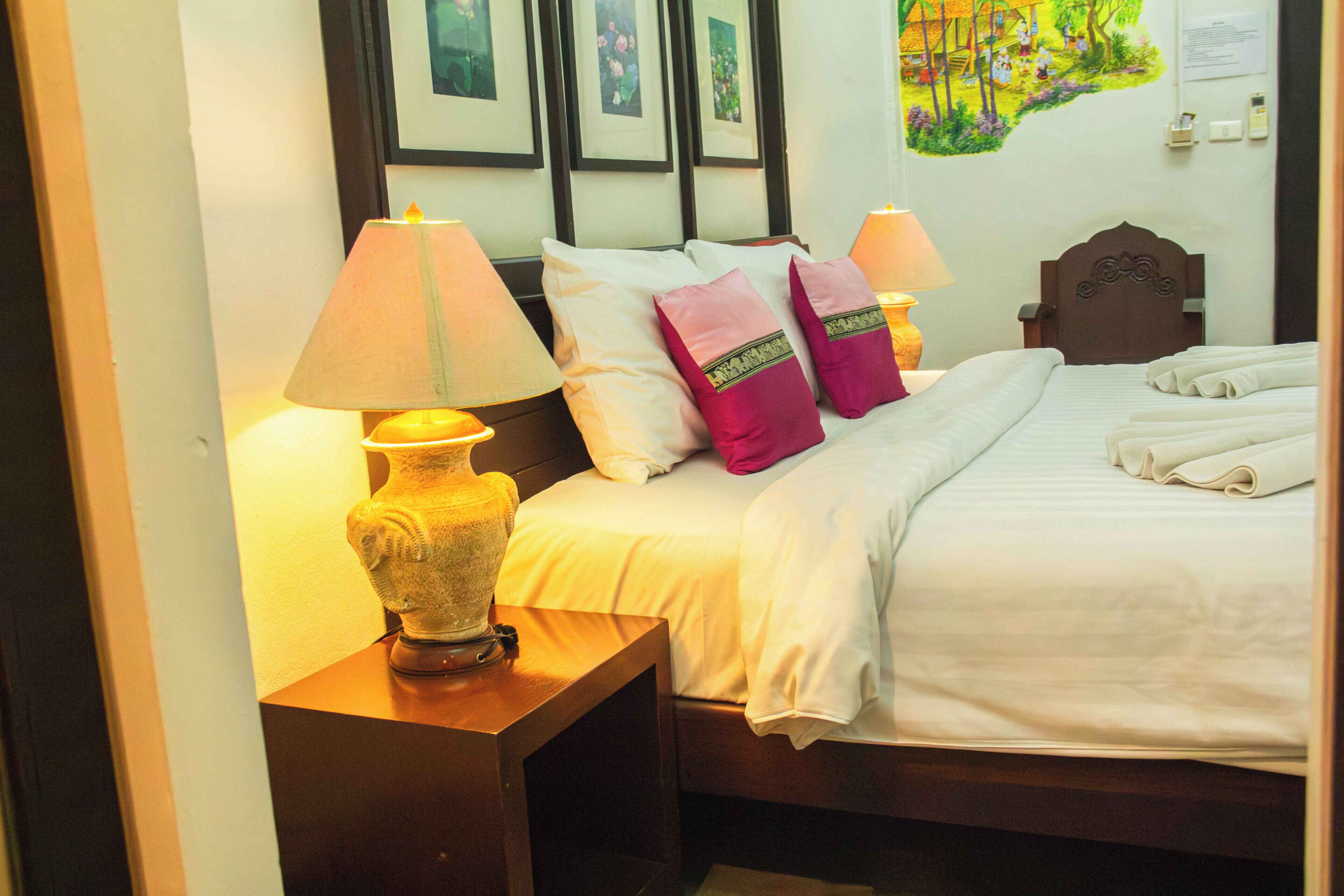 Una cama bien hecha con ropa blanca, dos almohadas con adornos, una mesita de noche de madera con una lámpara, y una pared con cuadros enmarcados.