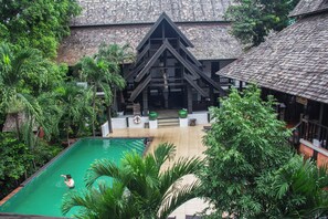 Zona de piscina con una persona nadando, rodeada de exuberante vegetación y un edificio con una estructura de techo triangular distintiva.