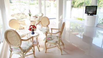 A bright, sunlit room with wicker furniture, a television, and a view of greenery outside.