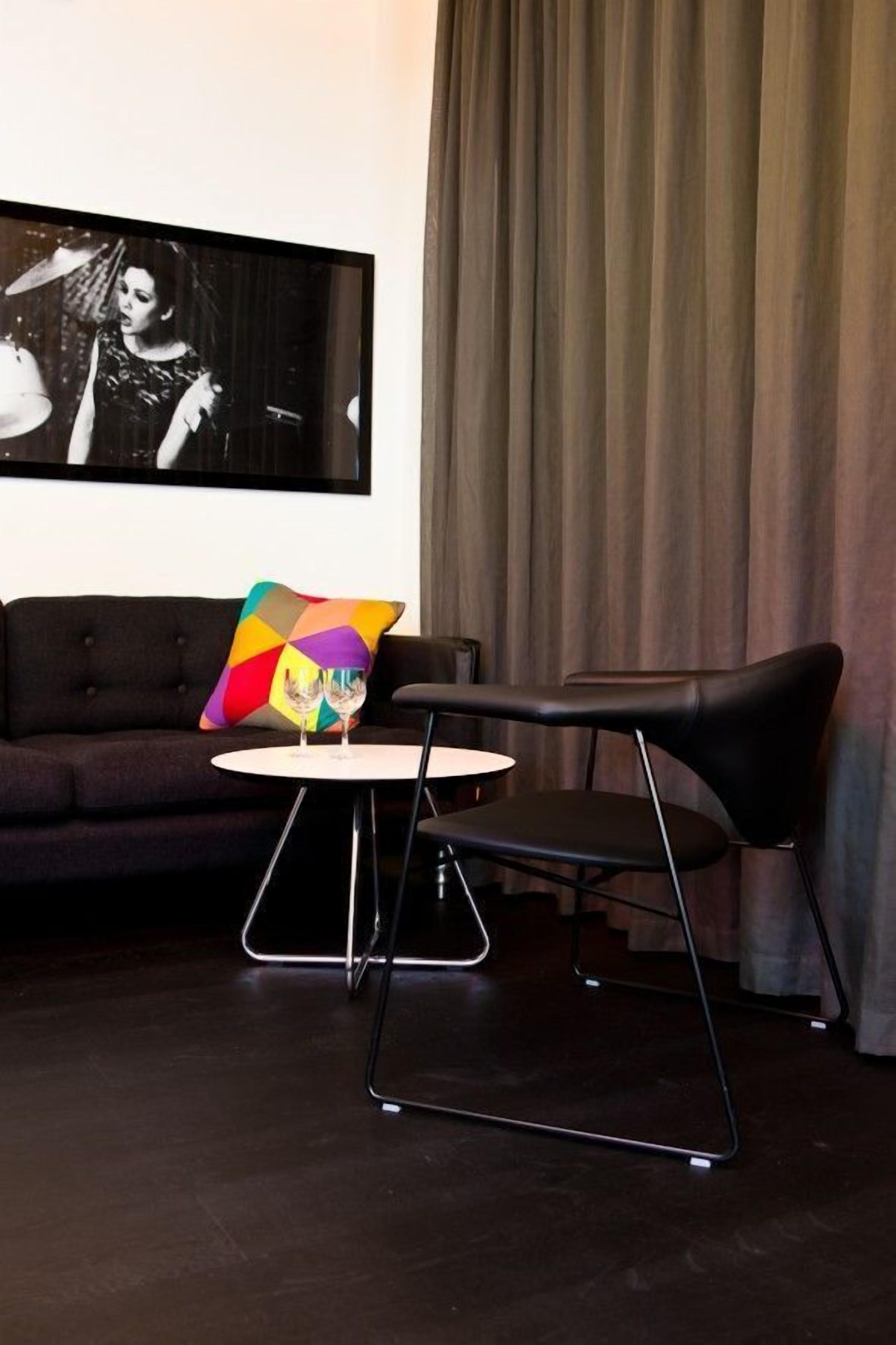 Una sala moderna con un sofá oscuro, una mesa redonda blanca y una silla negra. Un cojín colorido descansa sobre el sofá, y un cuadro enmarcado cuelga en la pared.