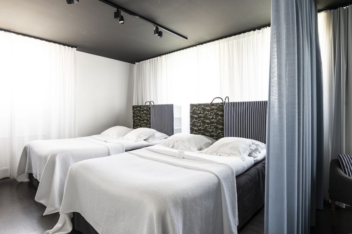 A modern hotel room with two single beds, a large window with sheer curtains, a dark ceiling, and a grey armchair.