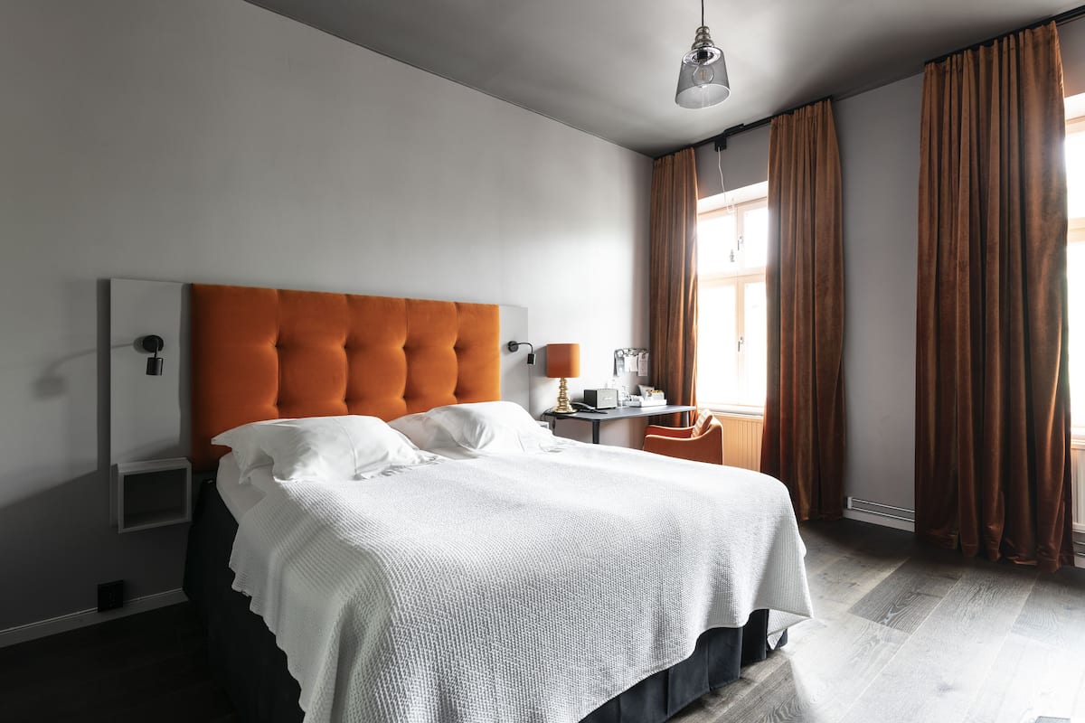 A modern hotel room with a large bed, a nightstand, a lamp, and a window with curtains.