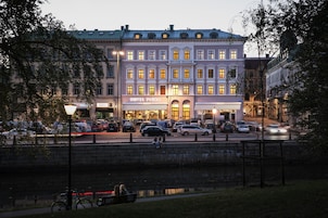 Hotel com fachada clássica, entrada visível e vista para a rua com carros estacionados e um canal.
