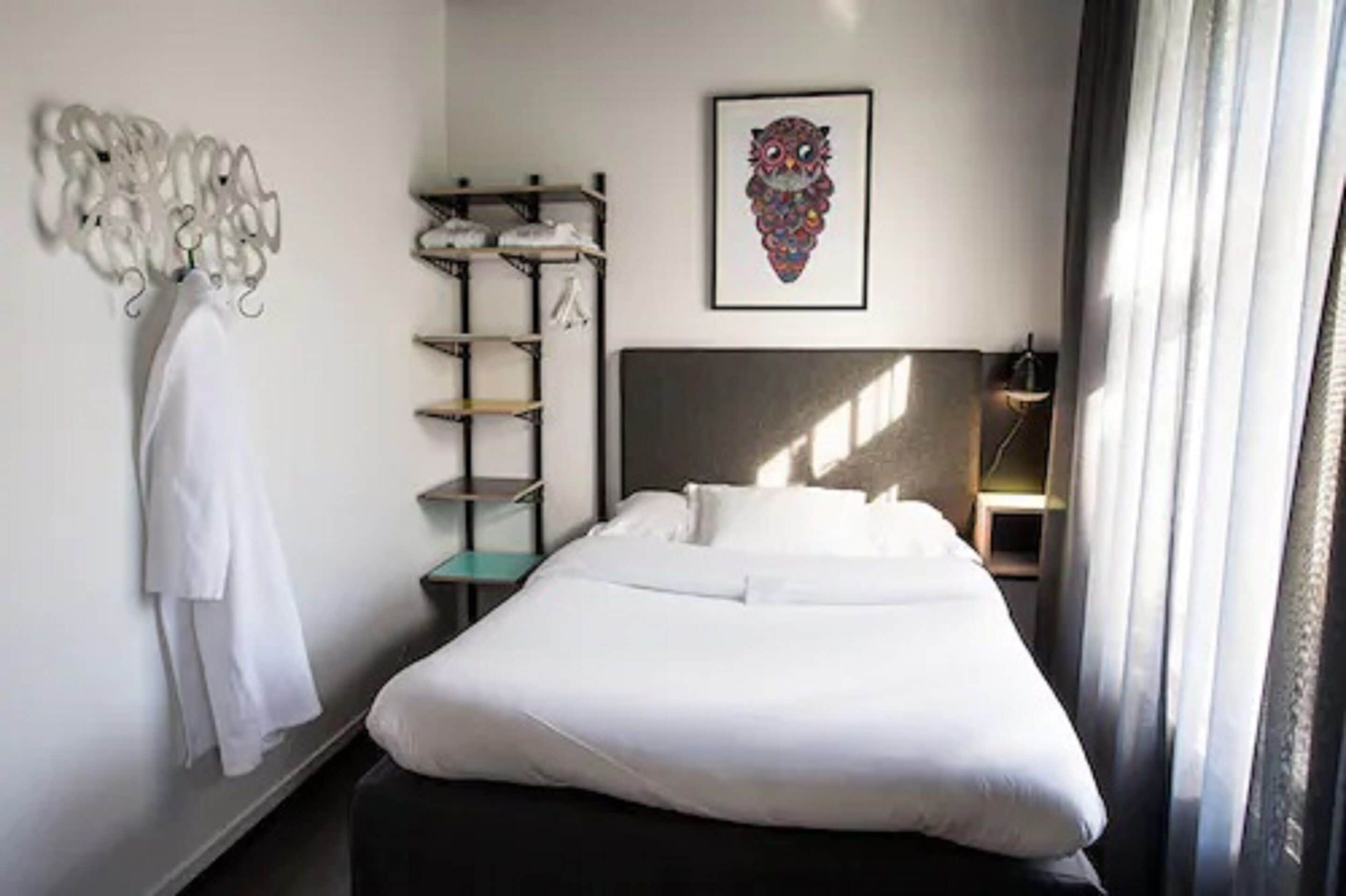 Una habitación de hotel con una cama bien hecha, un perchero en la pared, un estante con ropa doblada y una obra de arte enmarcada sobre la cama.
