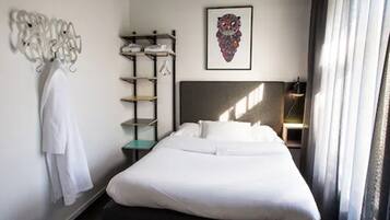 Una habitación de hotel con una cama bien hecha, un perchero en la pared, un estante con ropa doblada y una obra de arte enmarcada sobre la cama.