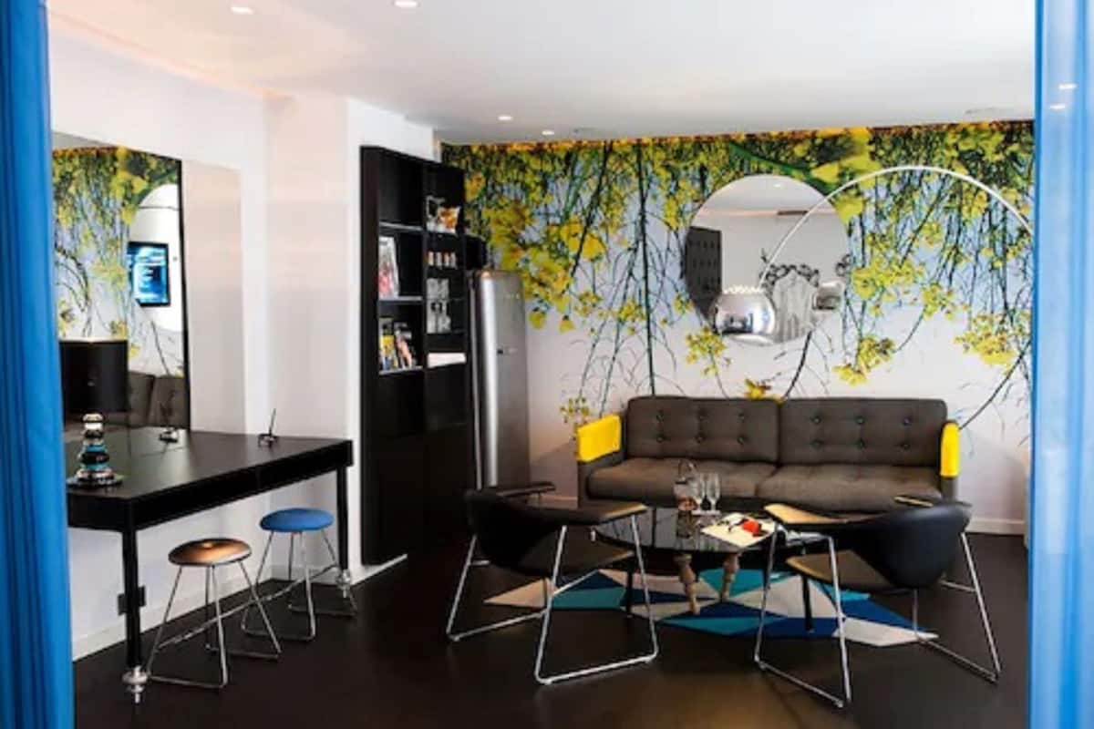A modern living room with a sofa, coffee table, and bar stools. There is a bookshelf and a refrigerator in the background.