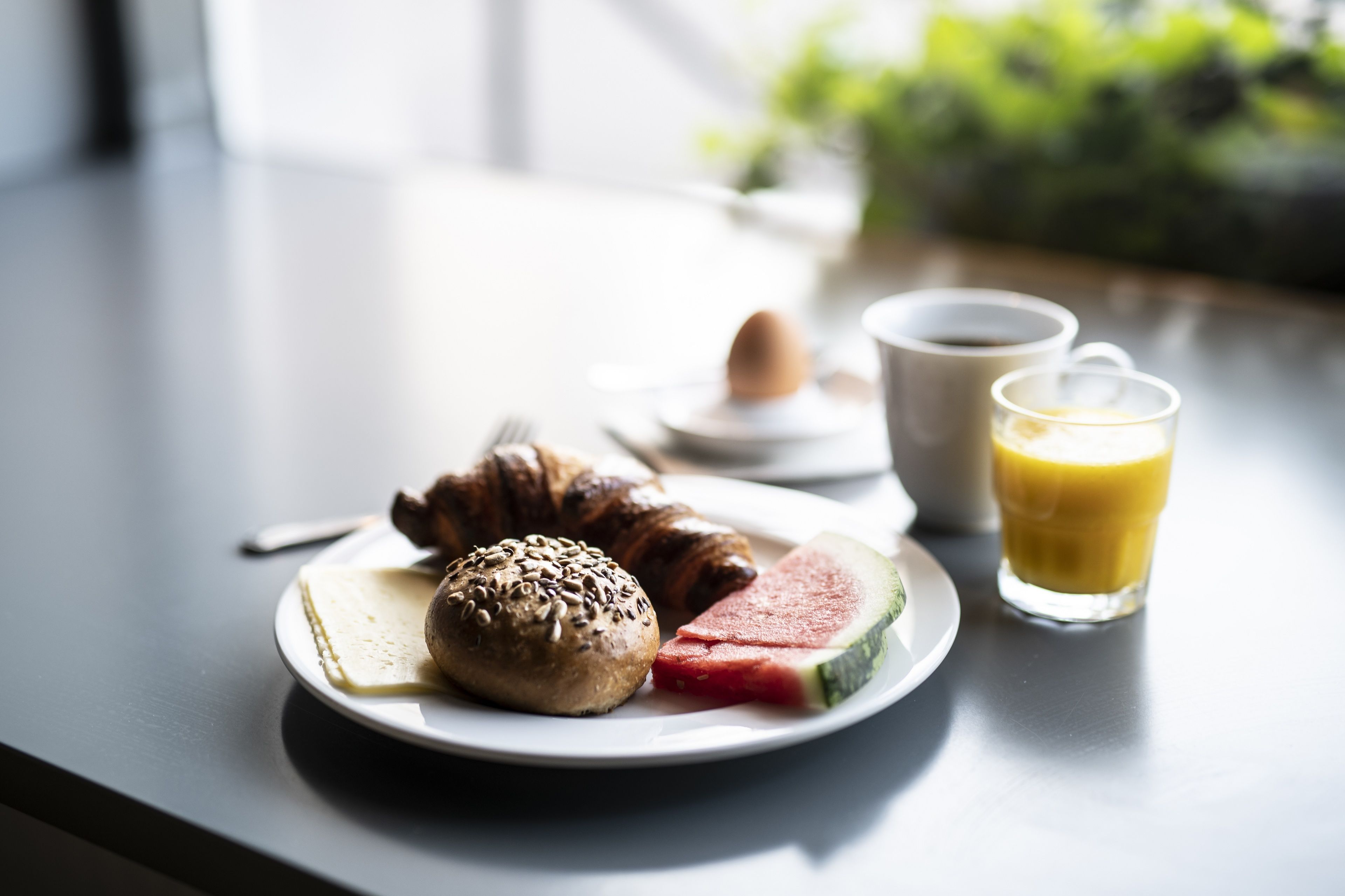 A breakfast plate with croissant, muffin, and watermelon slice, along with a cup of coffee and a glass of orange juice on a table.