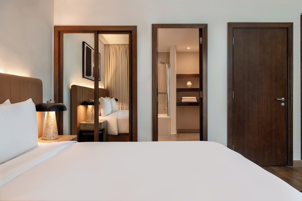 Habitación de hotel con cama, lámpara de noche, espejo, puerta y zona de ducha.