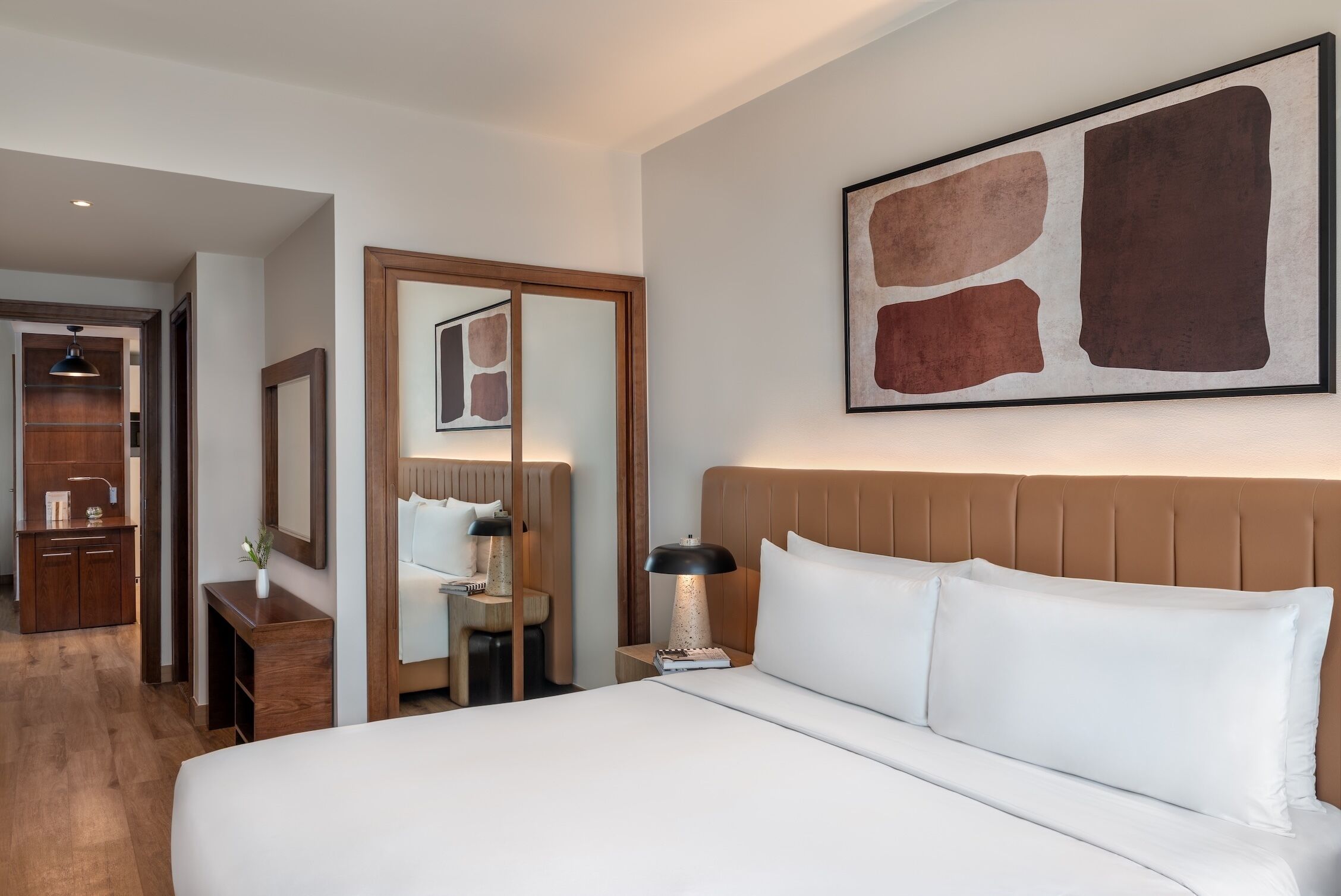 A hotel room with a large bed, a bedside table with a lamp, a mirror, and a wall-mounted abstract art piece.
