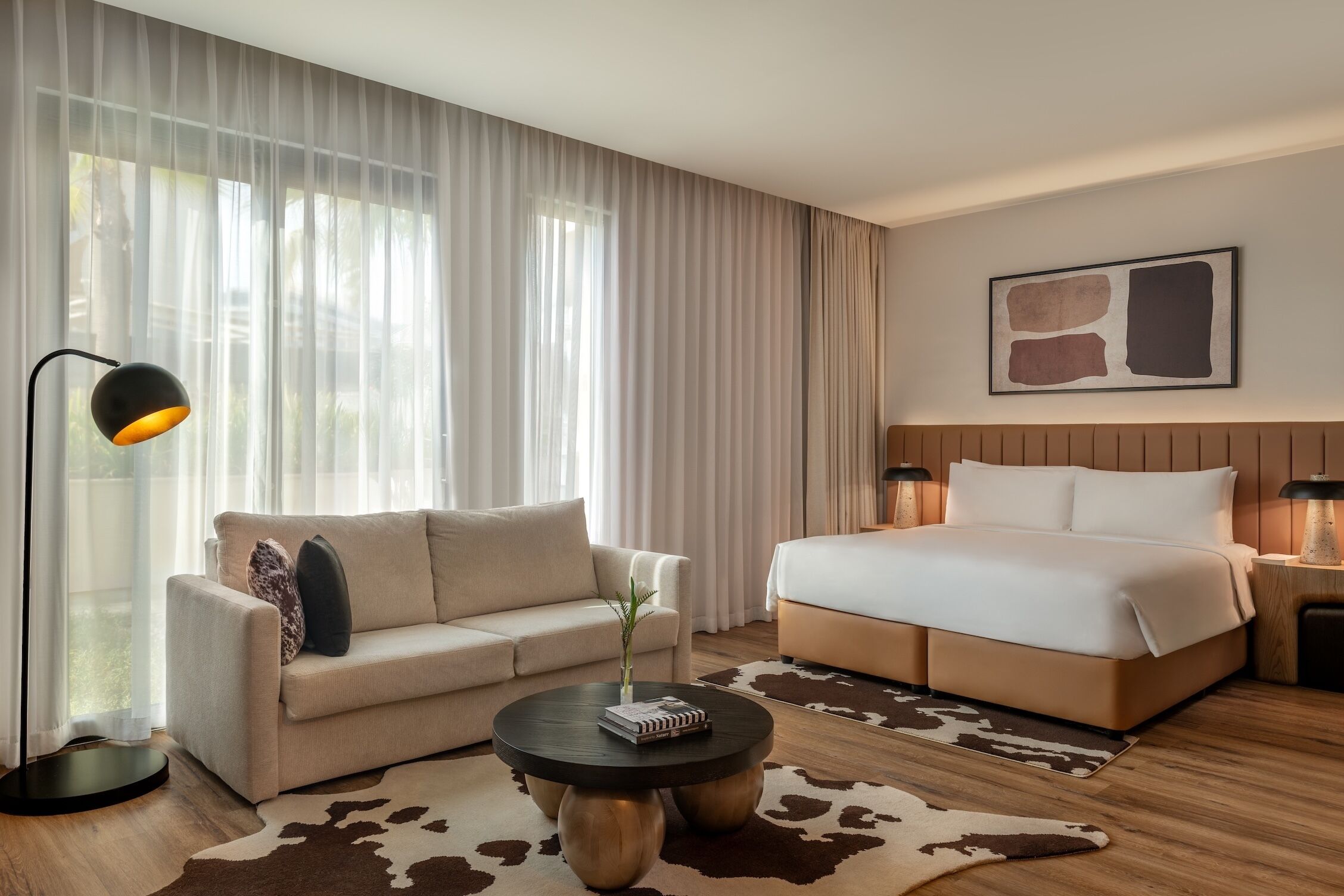 A modern hotel room with a sofa, a round coffee table, a bed, and a large window with sheer curtains.