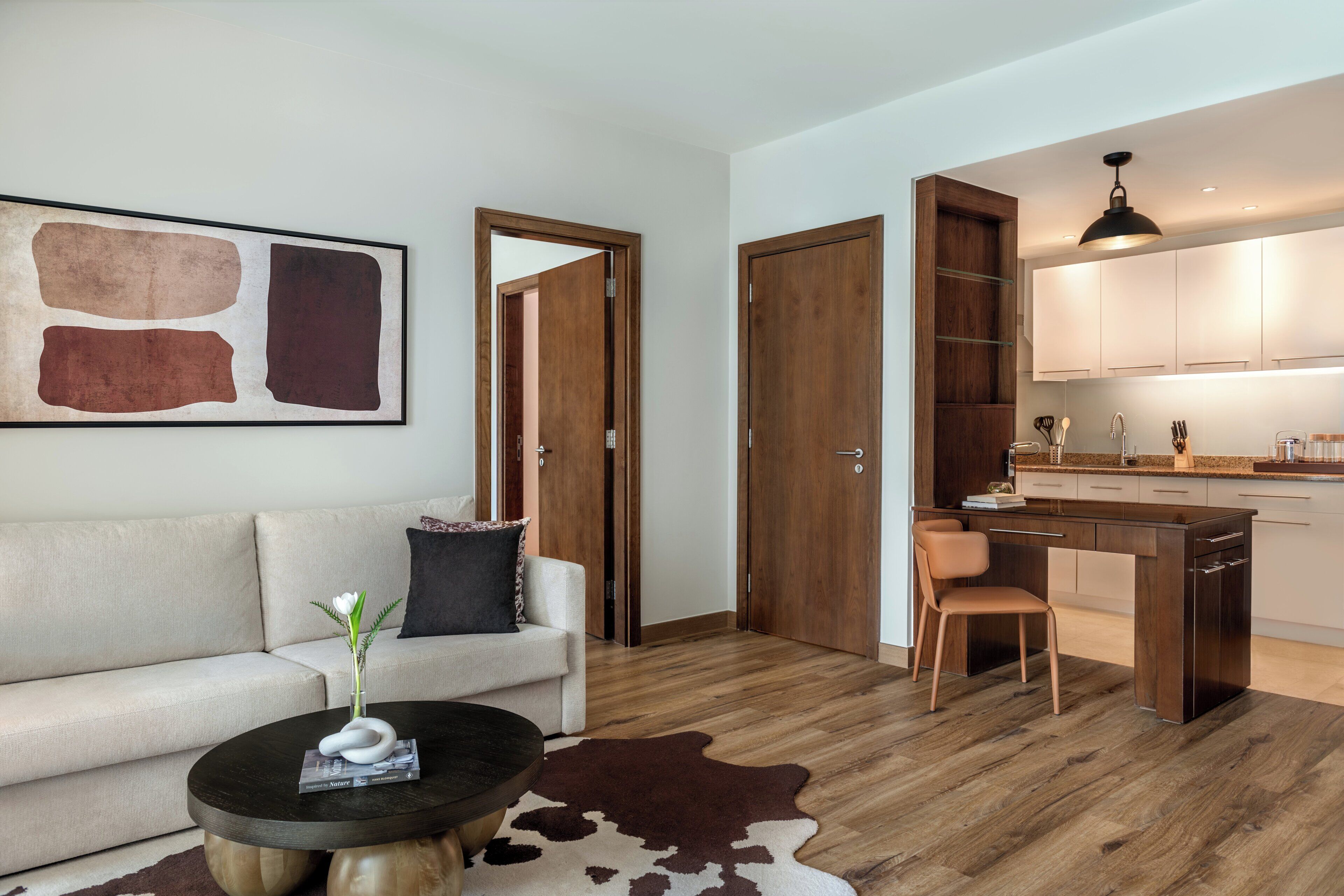 A modern living room with a sofa, coffee table, and a painting. There is a kitchen area with wooden cabinets and a dining table.