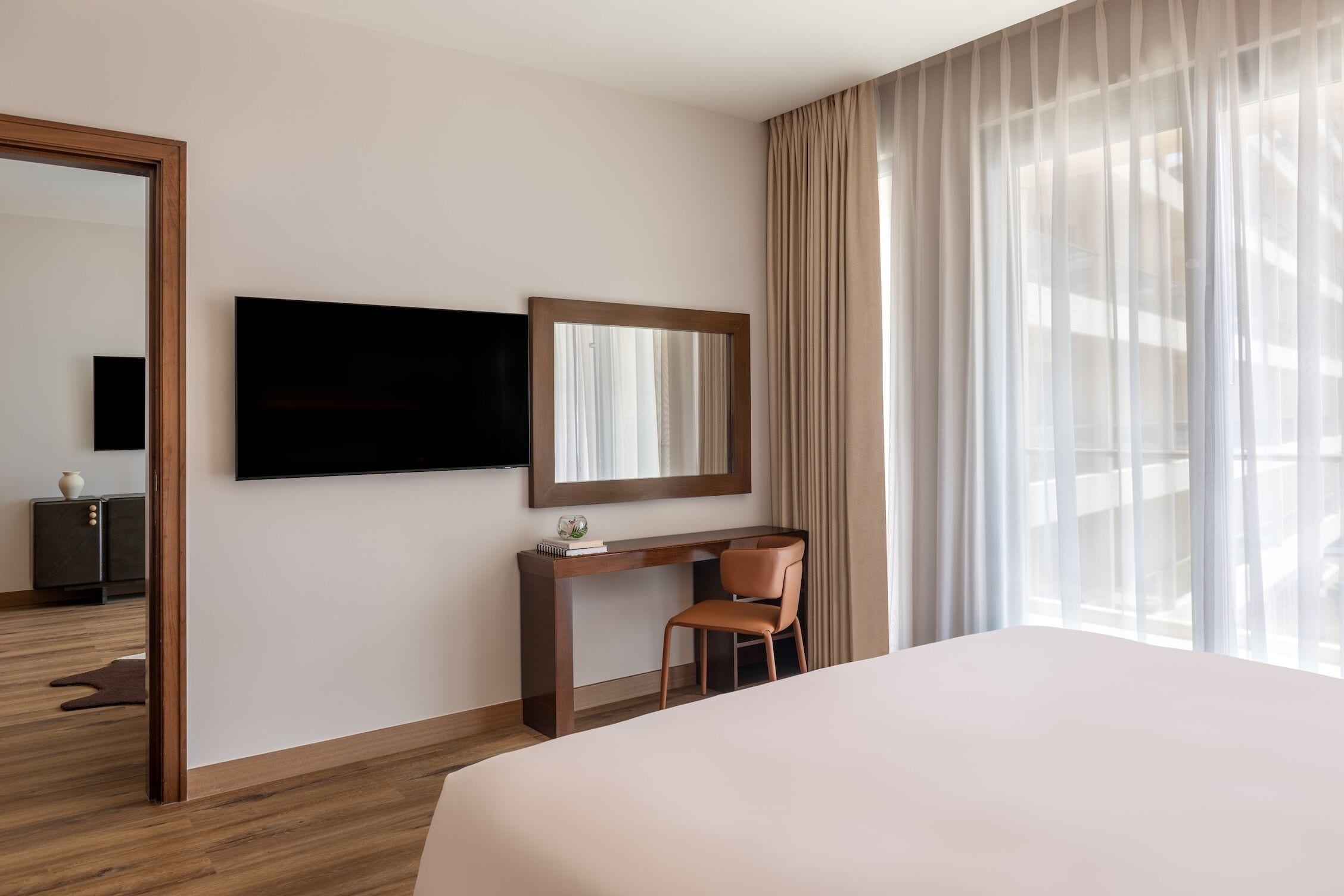 A hotel room with a bed, a desk with a chair, a TV mounted on the wall, and a large window with sheer curtains.