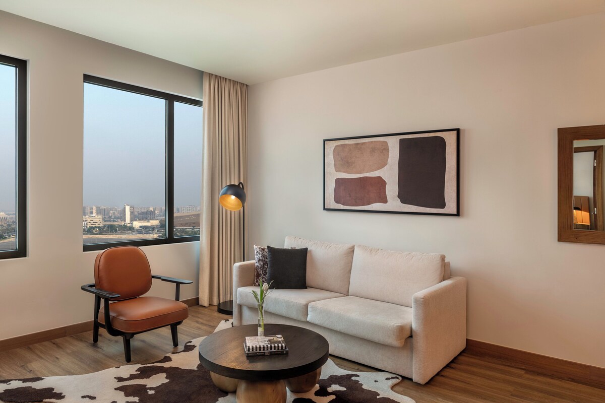 Una habitación de hotel moderna con un ventanal grande, un sofá, una silla, una mesita de centro y un cuadro en la pared.