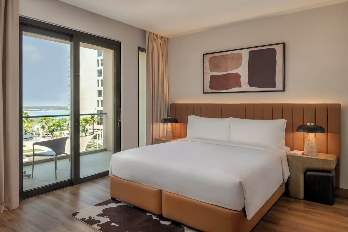 Ein Hotelzimmer mit einem großen Bett, zwei Nachttischlampen, Blick aufs Meer und einem modernen abstrakten Gemälde an der Wand.