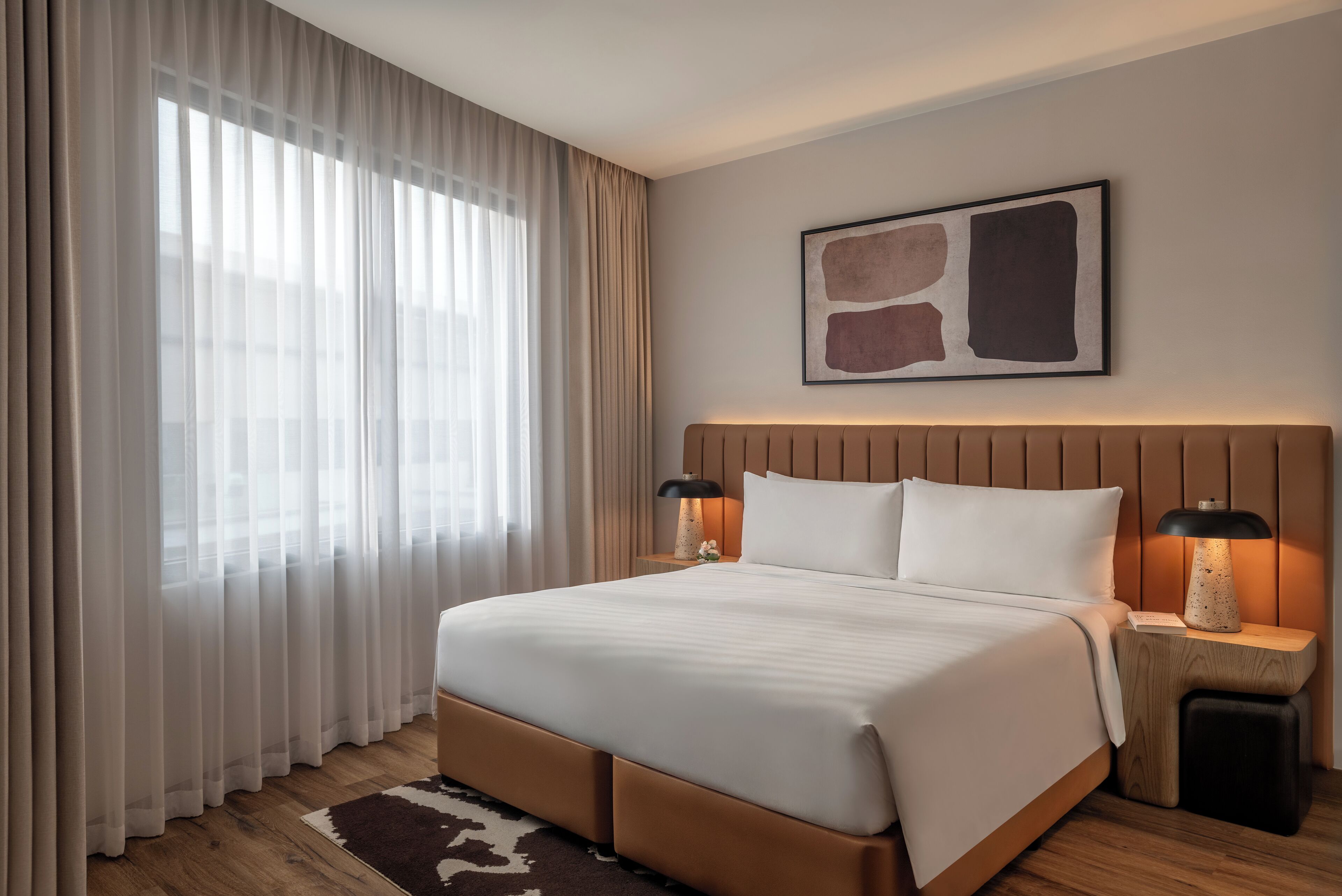 Habitación de hotel con una cama grande, dos mesitas de noche con lámparas, una obra de arte colgada en la pared y cortinas de gasa.