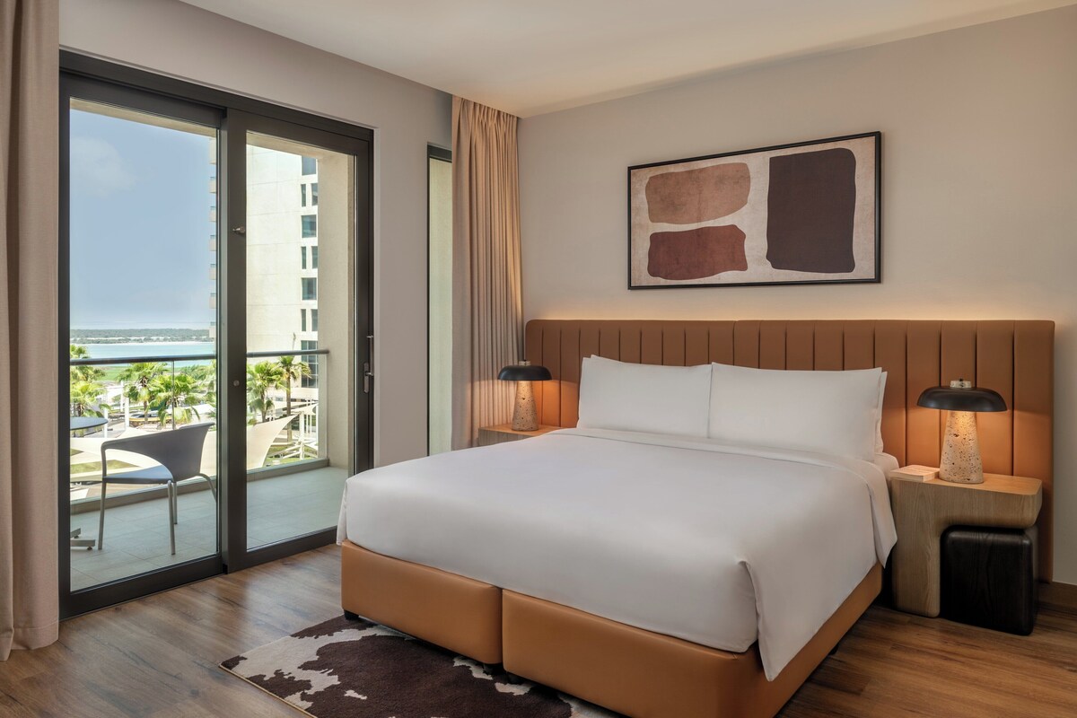 Habitación de hotel con una cama grande, dos lámparas de noche, vista al océano y un cuadro abstracto moderno en la pared.