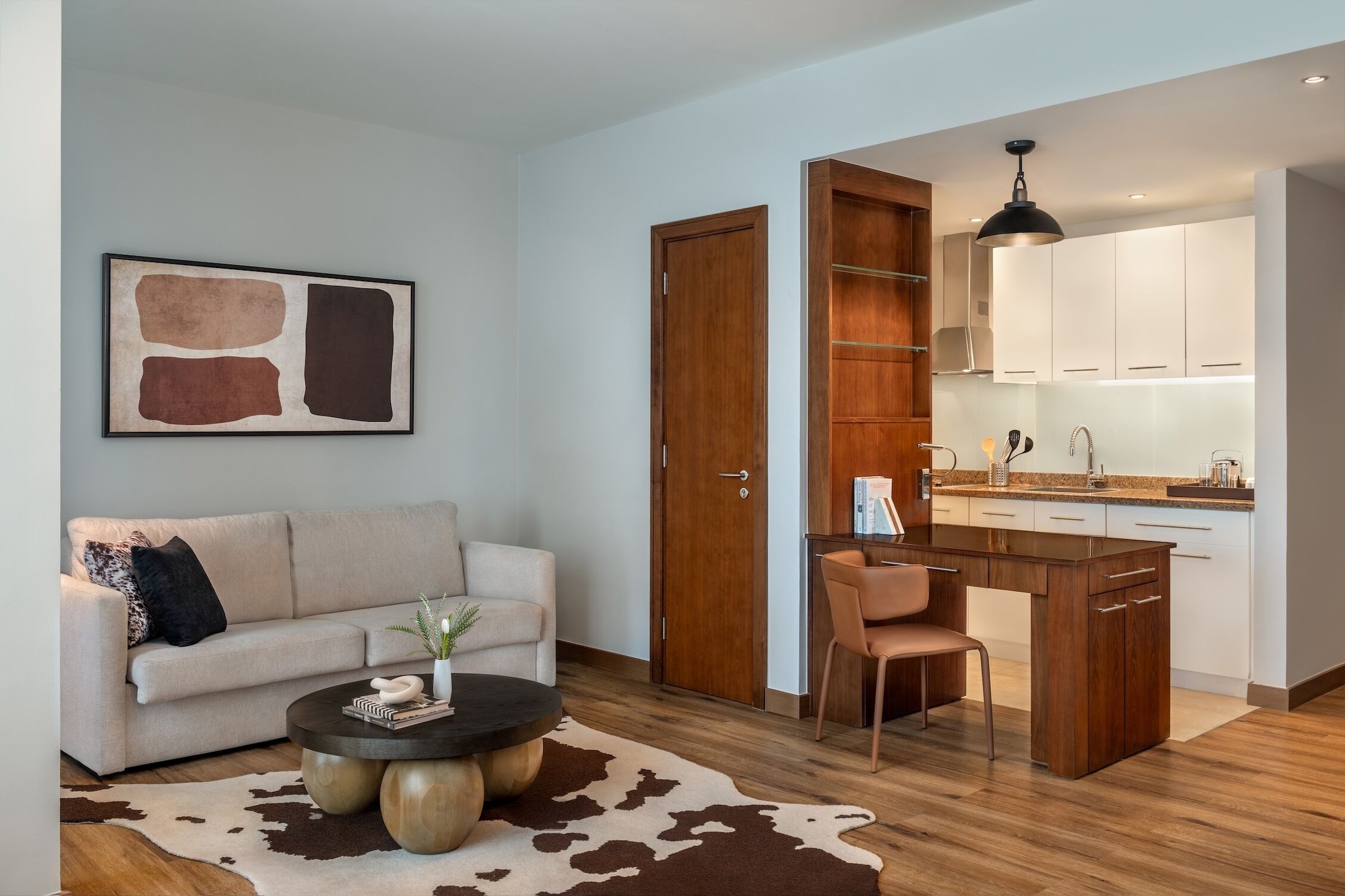 A modern living room with a sofa, a coffee table, and a cowhide rug. Adjacent is a kitchen with wooden cabinets and a pendant light.