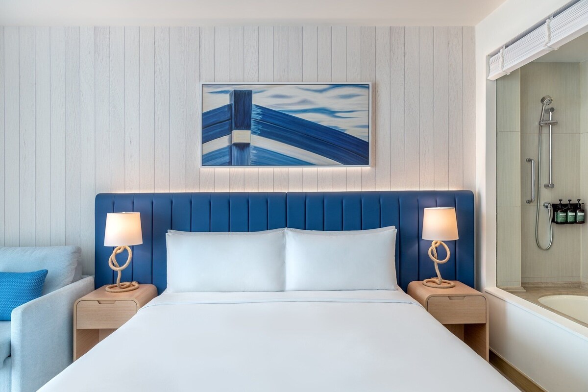 A hotel room with a large bed, two bedside tables with lamps, a headboard, a painting on the wall, and a shower area.