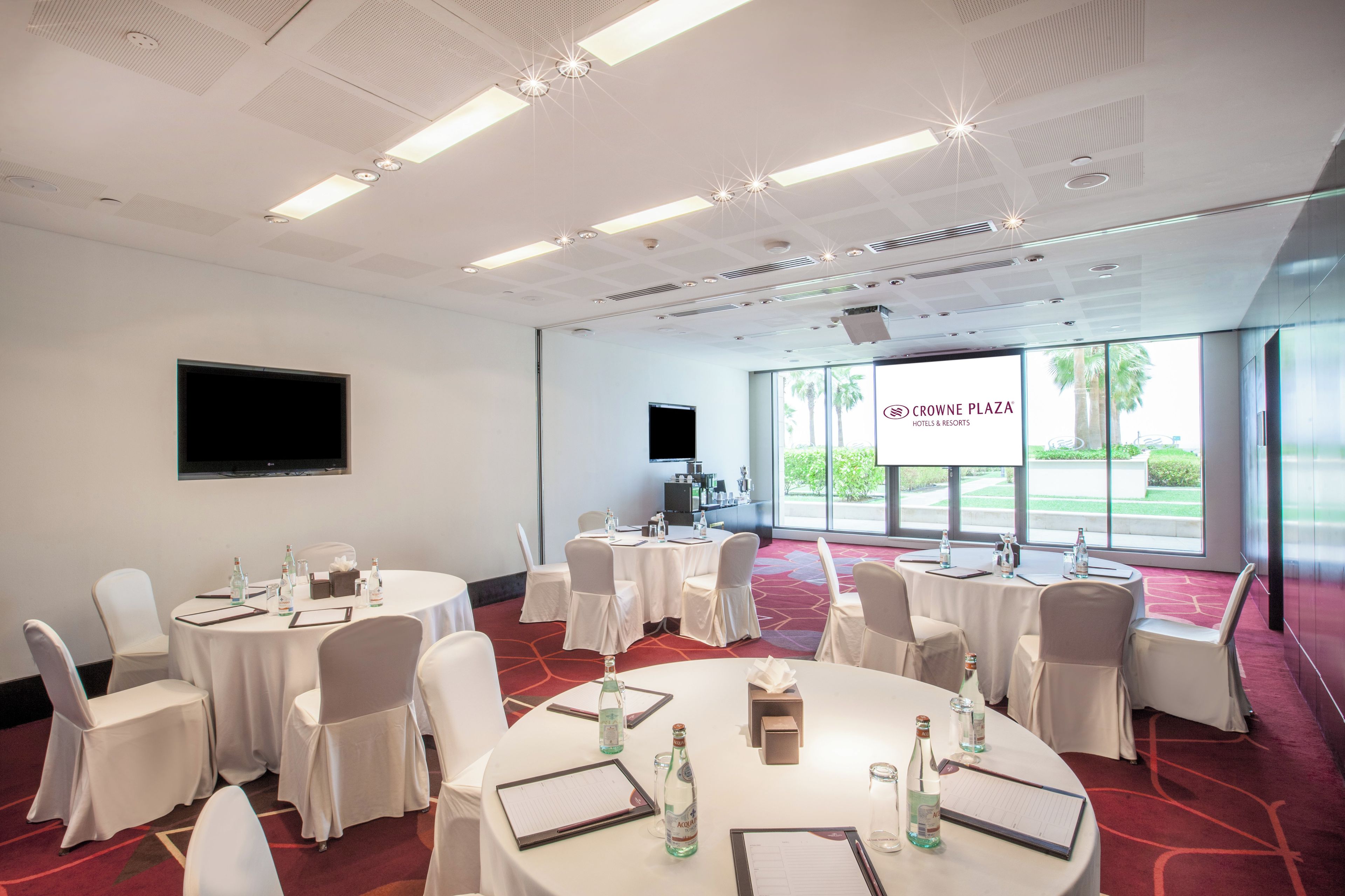 Een congreszaal met meerdere tafels, stoelen en een groot scherm waarop 'CROWNE PLAZA HOTELS & RESORTS' staat.