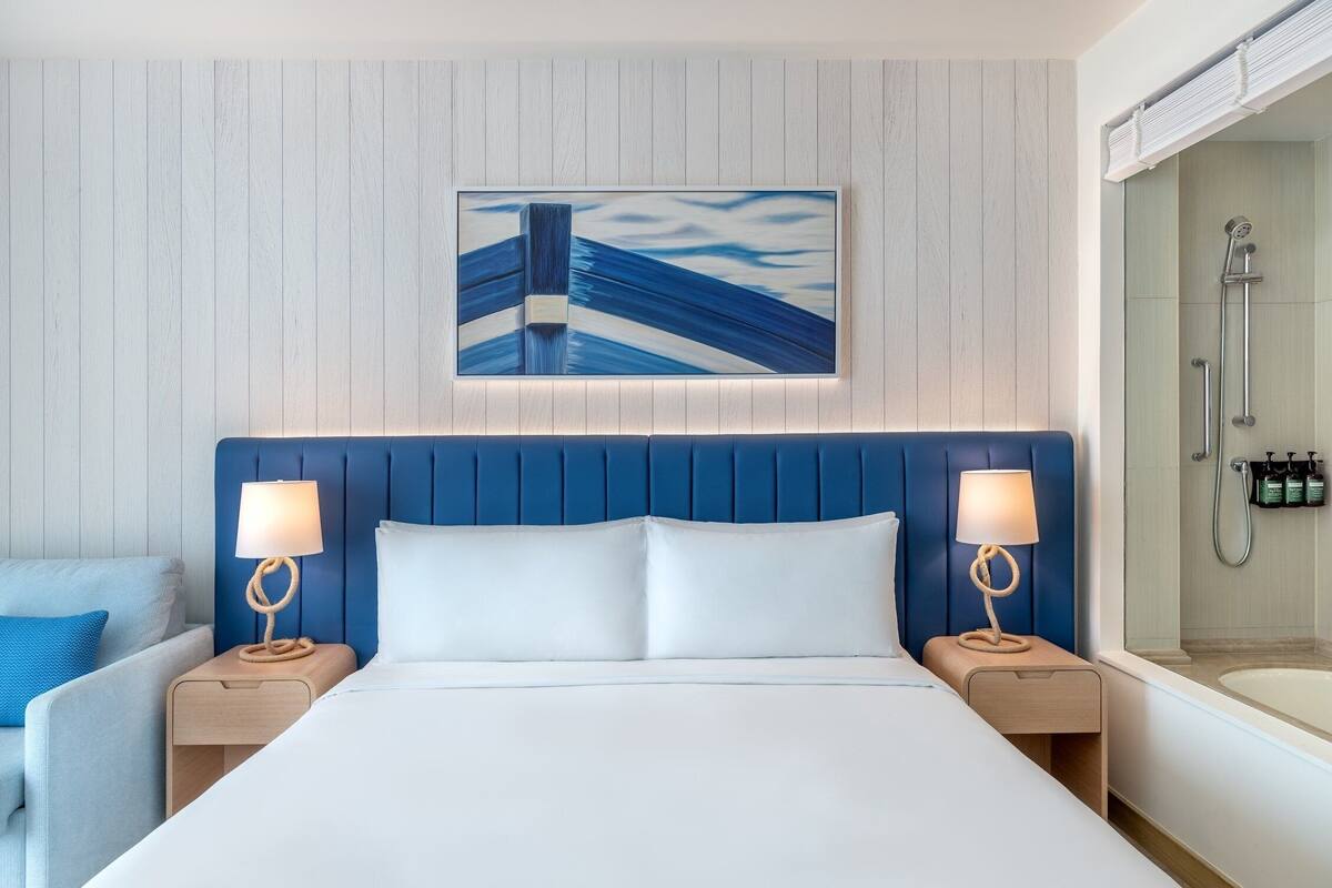 A hotel room with a large bed, two bedside tables with lamps, a headboard, a painting on the wall, and a shower area.