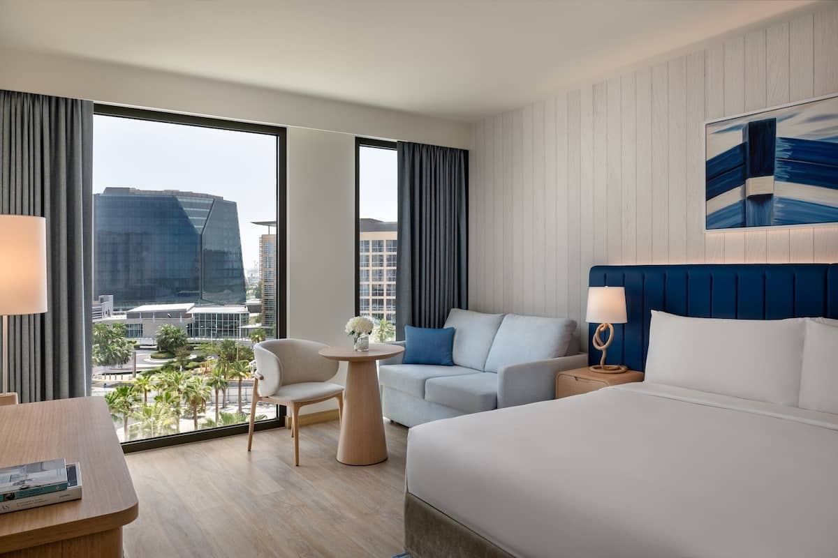 A modern hotel room with a large bed, a sofa, a round table, and a view of the cityscape.