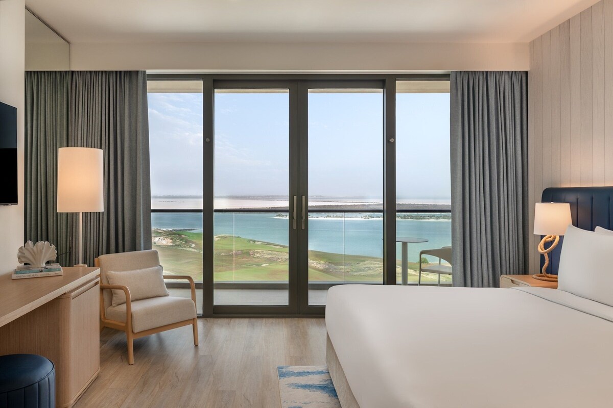 A hotel room with a large sliding glass door to the sea, a bed, a desk, and a chair.
