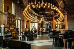 A spacious restaurant with multiple tables and chairs, a central hanging light fixture, and a large window providing natural light.