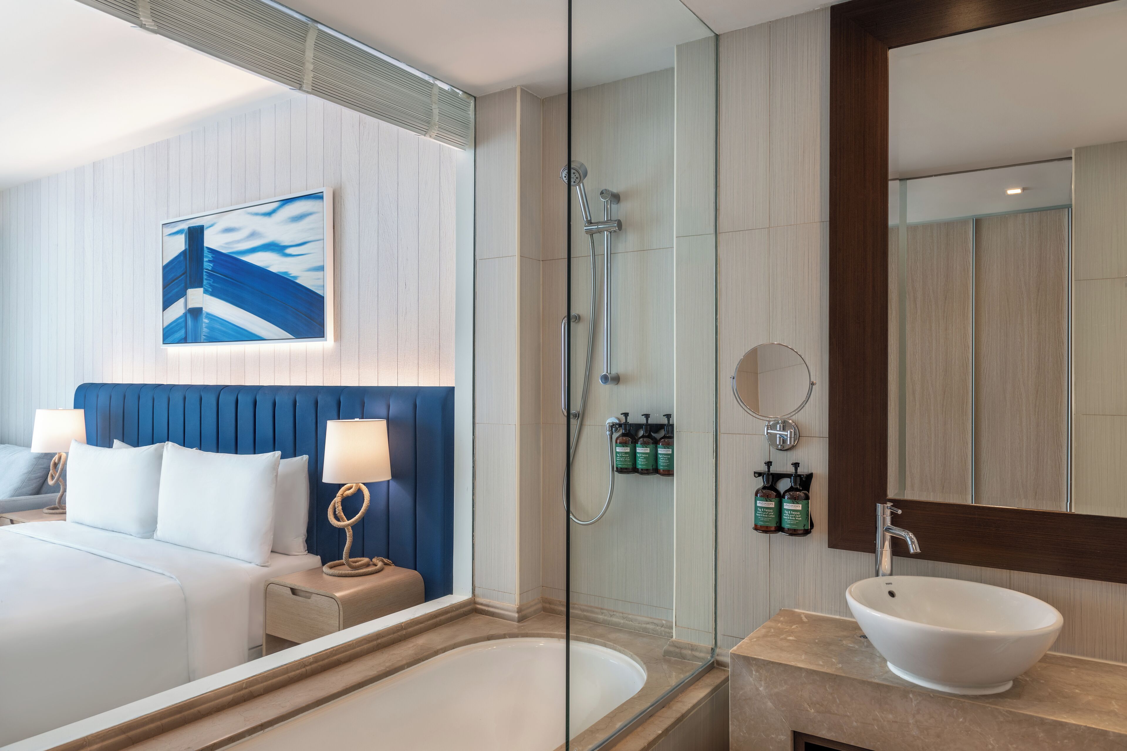 A modern hotel bathroom with a glass-enclosed shower, a large bed with a blue headboard, and a white sink.