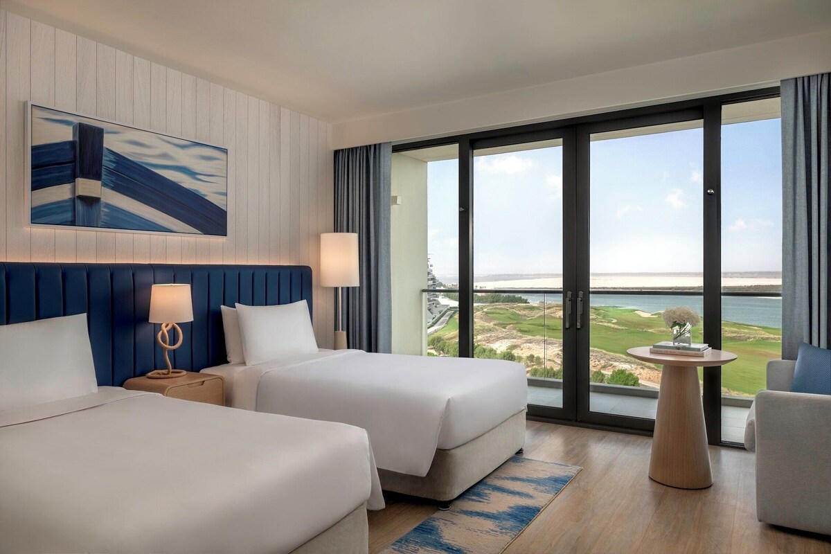 A hotel room with two beds, a large window with a view of a golf course, and a small table with a lamp.