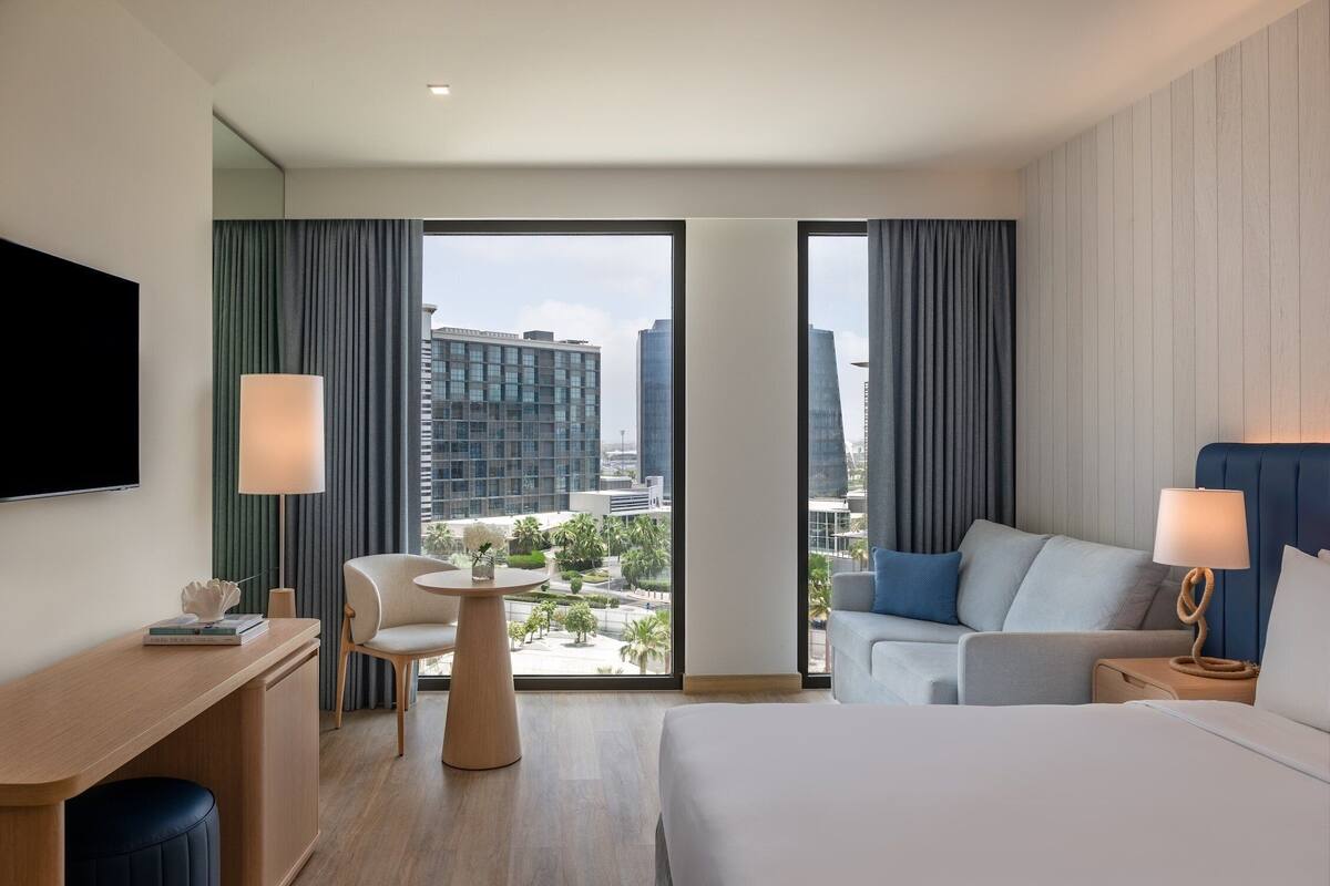 A modern hotel room with a large bed, a desk, a chair, a sofa, and a view of the cityscape.