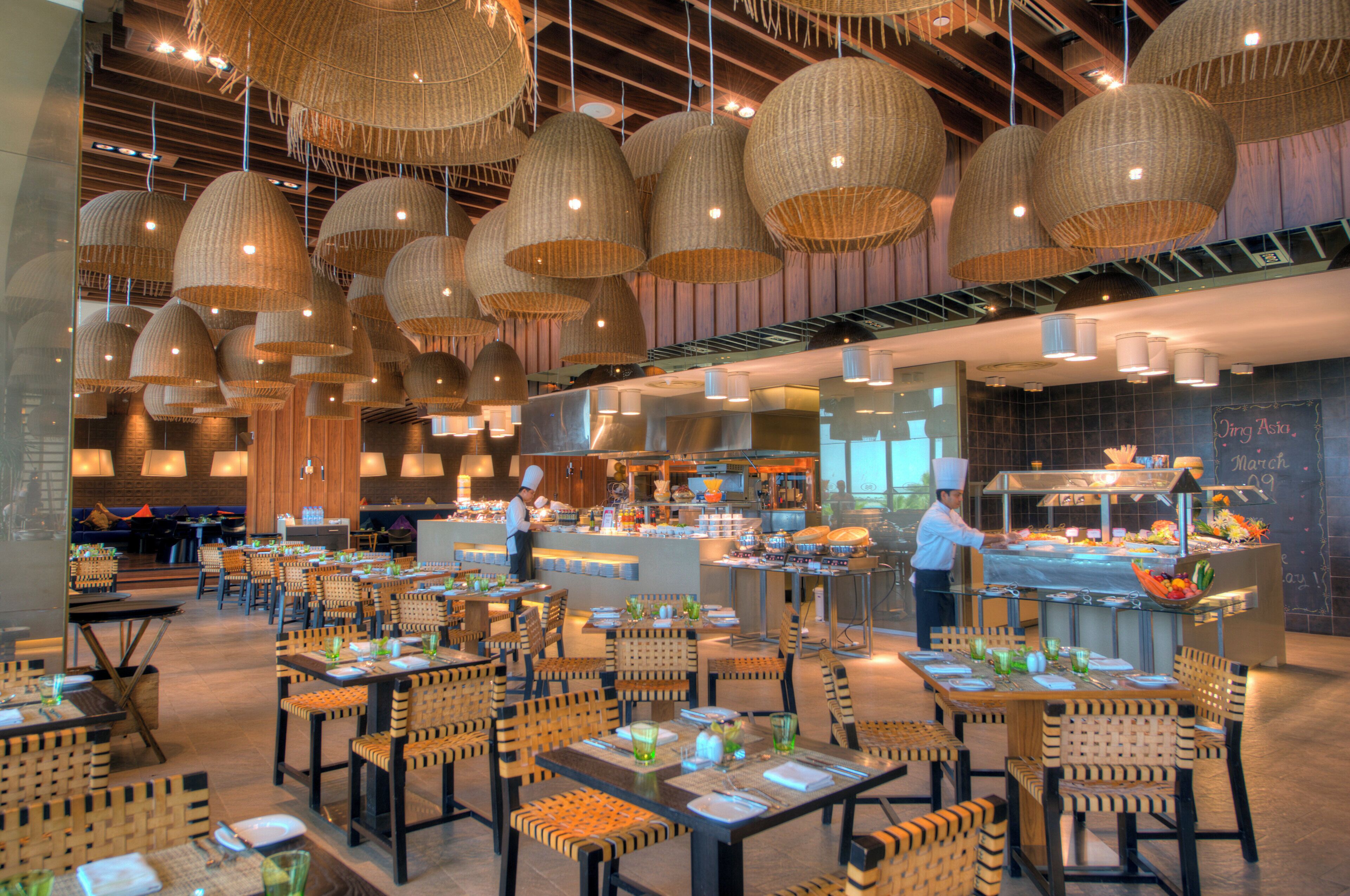 A restaurant interior with multiple dining tables and chairs, a kitchen area, and hanging wicker lamps.