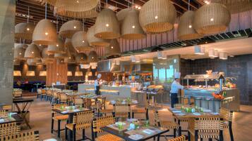 A restaurant interior with multiple dining tables and chairs, a kitchen area, and hanging wicker lamps.