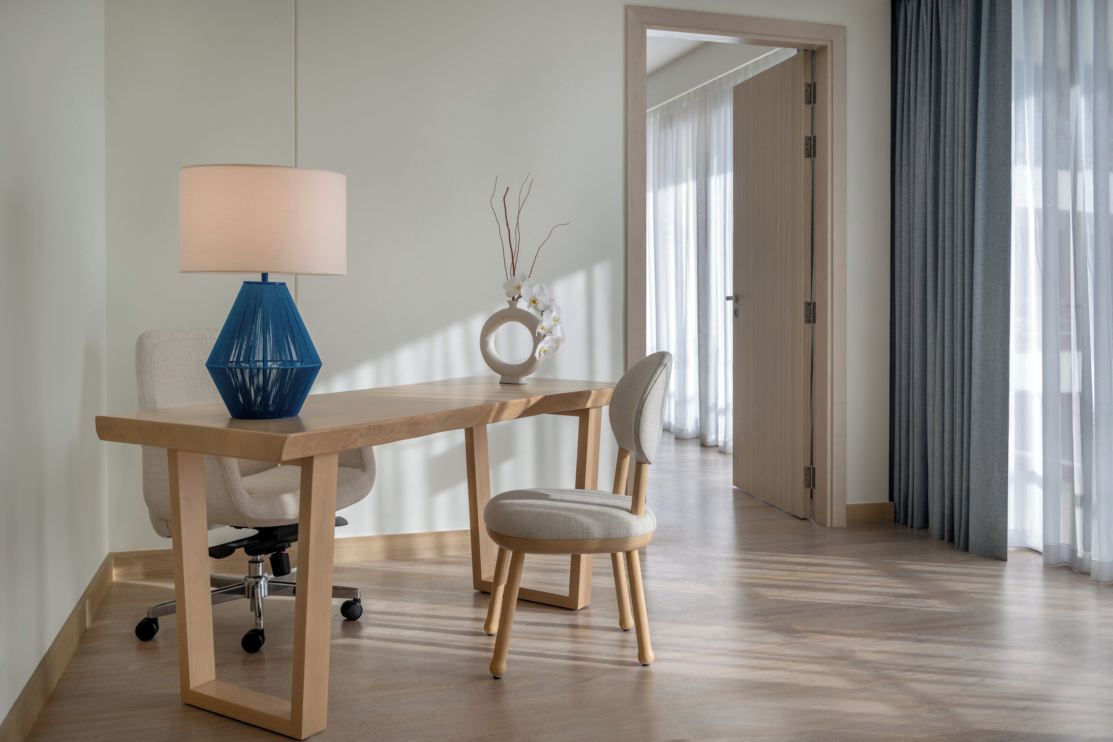 A modern room with a wooden desk, a blue lamp, a white chair, and a vase with white flowers.