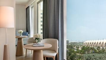 A modern hotel room with a large window, a round table with a book and flowers, a sofa, and a view of a unique building and palm trees.