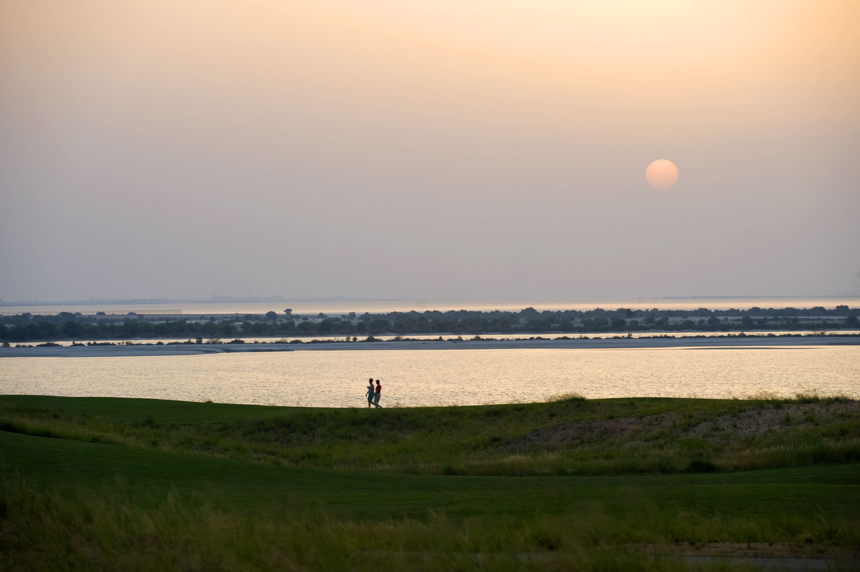 Een rivier bij zonsondergang, met twee personen die over een grasachtige oever wandelen.