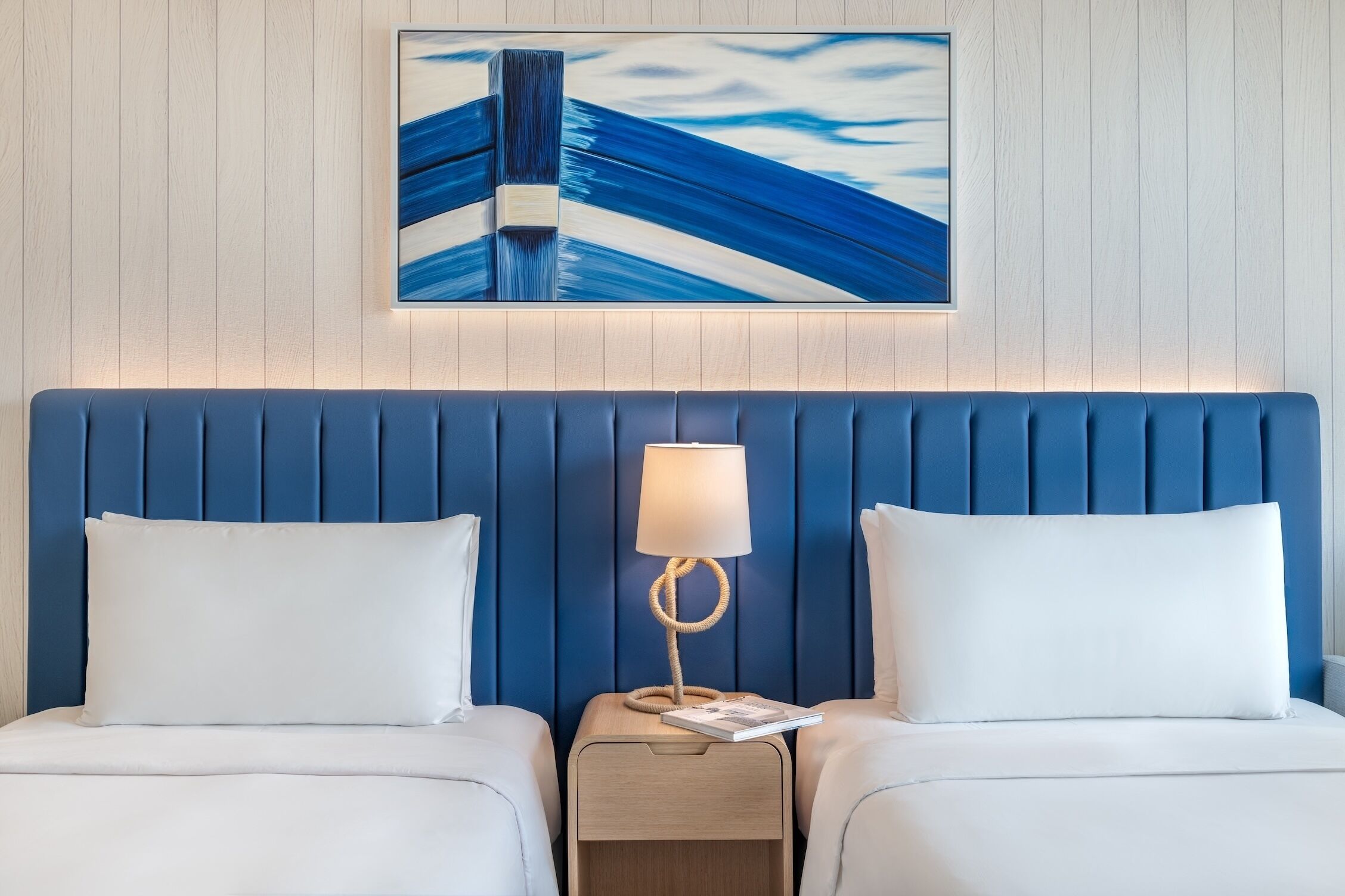 A hotel room with a blue headboard, two white beds, a bedside table with a lamp, and a framed artwork above the headboard.