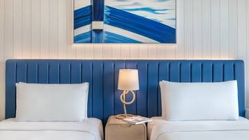 A hotel room with a blue headboard, two white beds, a bedside table with a lamp, and a framed artwork above the headboard.