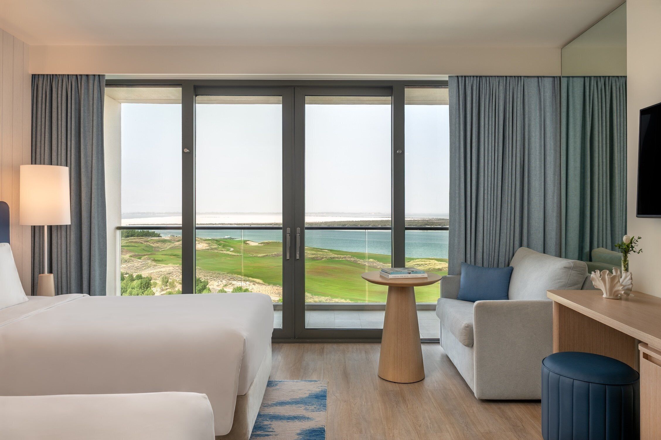 A hotel room with a large window, a bed, a sofa, a small table, and a TV.